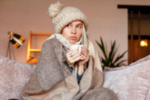 A woman sitting on a couch with a mug, a knit hat, and a blanket wrapped around her.